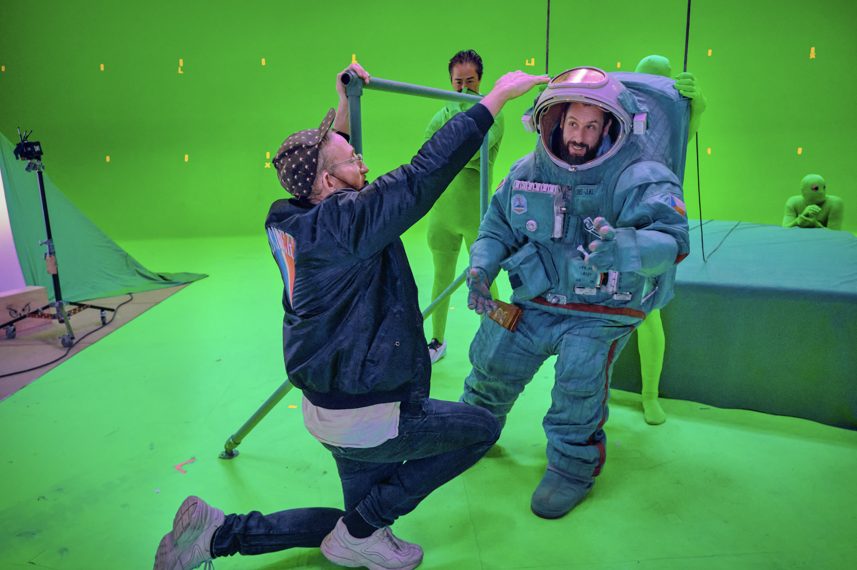 An actor in a spacesuit looks off from the camera in a film studio wrapped with a green screen; a person in a jean jacket adjusts his suit, and in the background, people wearing green screen bodysuits adjust the space